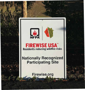 White street sign leaning against a tree trunk declaring the location as a "NFPA Firewise USA Nationally Recognized Participating Site."