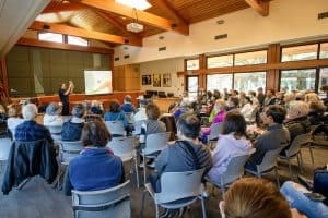 A large group in a community room listens to a presentation. The presenter is holding up a booklet.